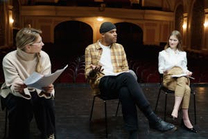 Three actors rehearsing on a theater stage, discussing scripts and performances.