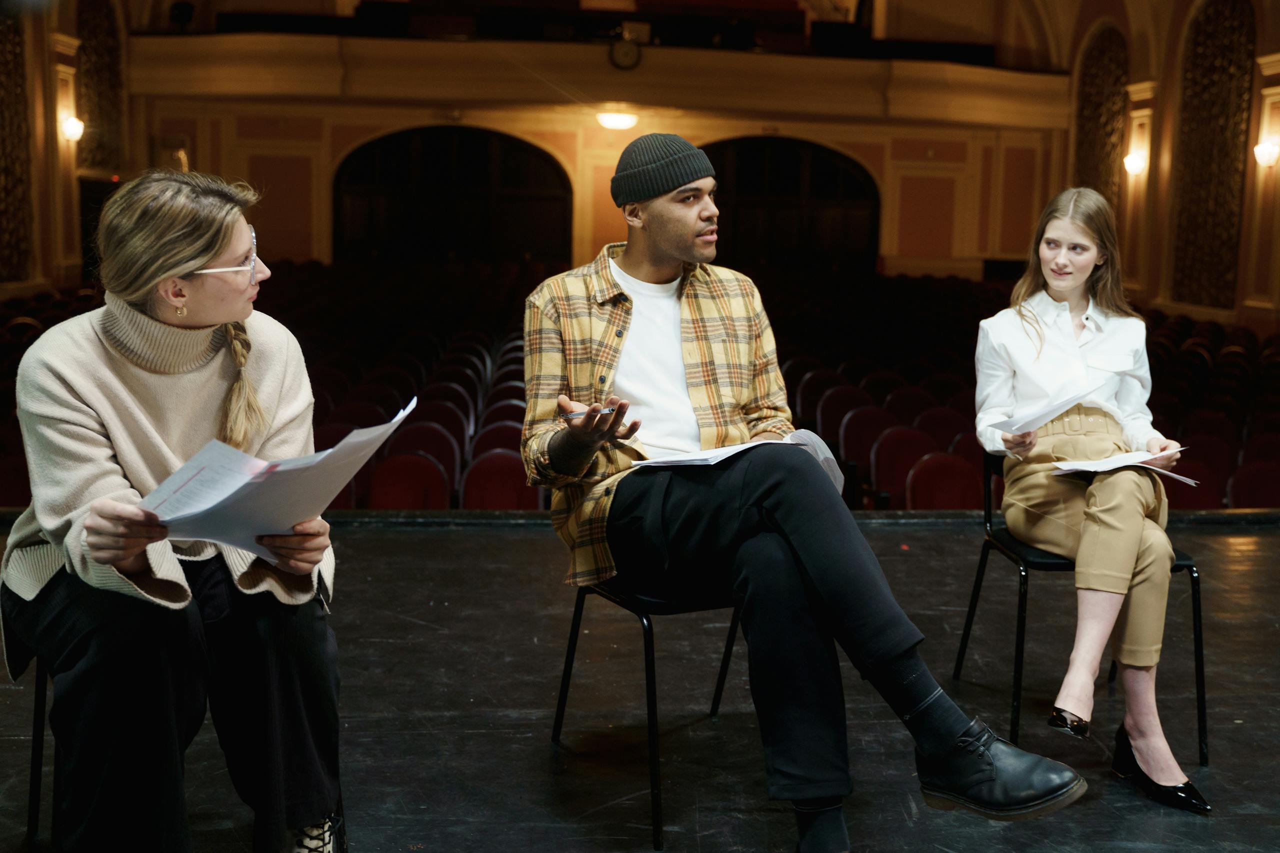 Three actors rehearsing on a theater stage, discussing scripts and performances.